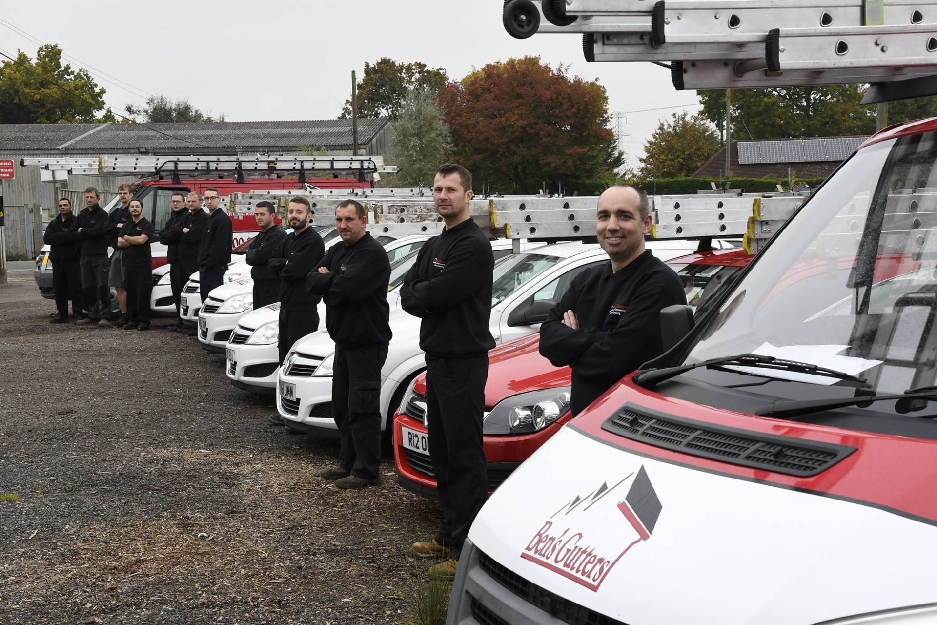 Line of white vans and drivers standing beside them at an outdoor event on a gravel lot