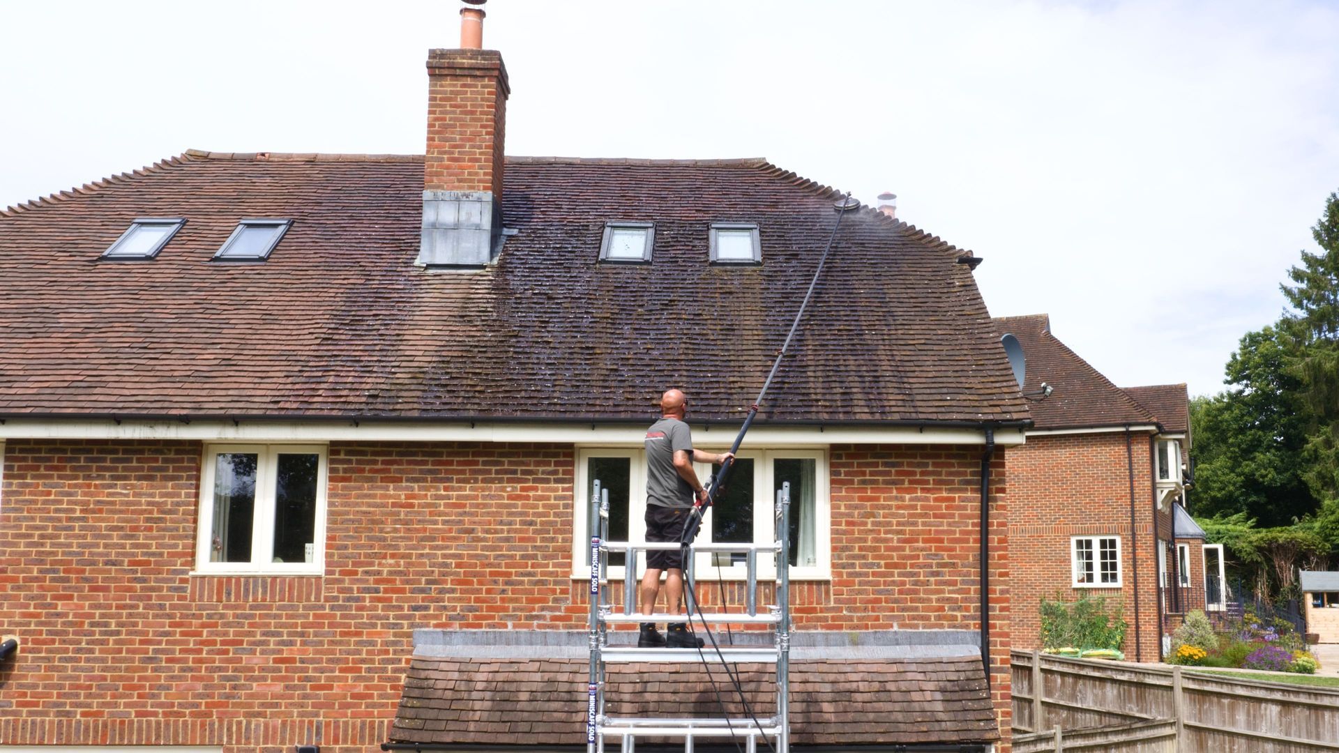 Worker on scaffolding painting a brick house’s exterior wall under a tiled roof
