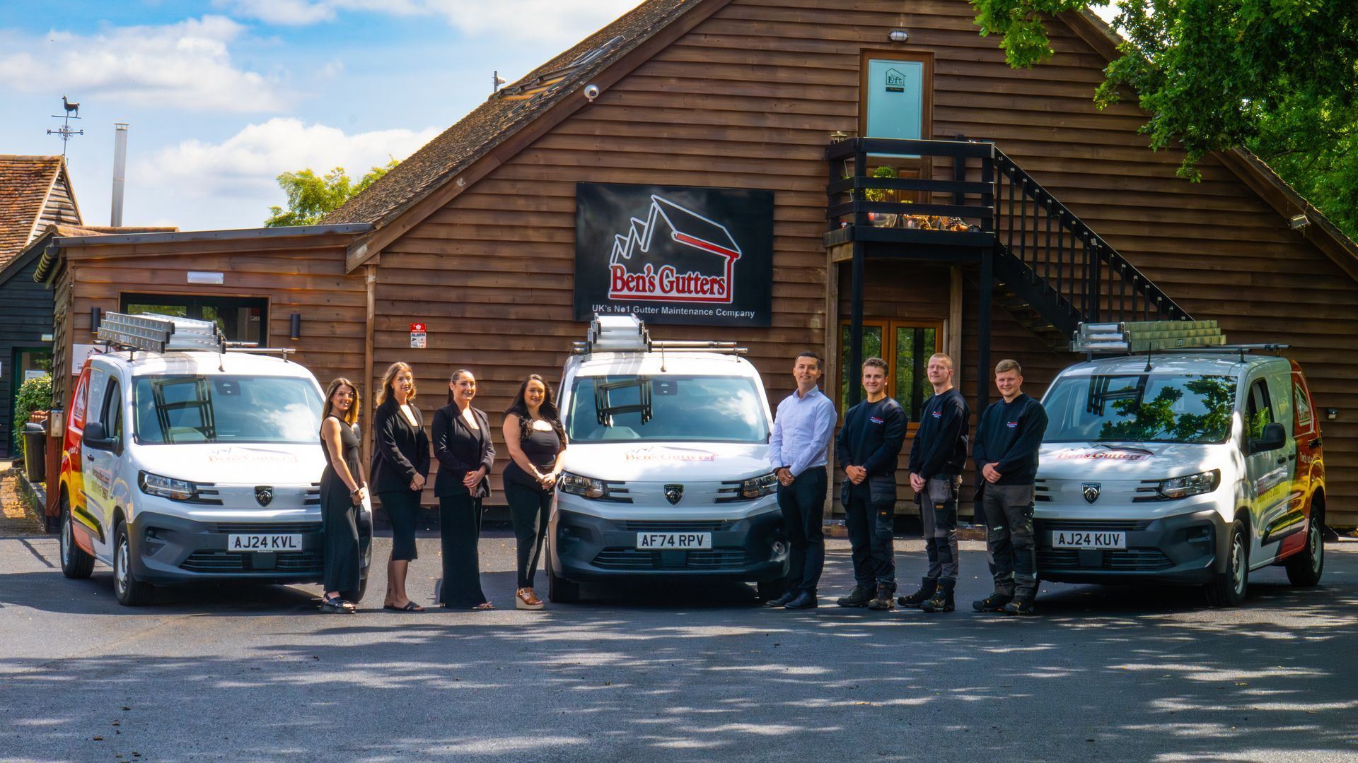 Group posing with white vans outside a wooden building, likely a business or fleet yard.