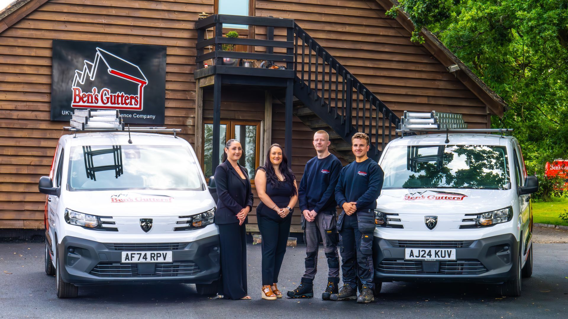 Four people standing between two white vans outside a wooden lodge with a sign overhead.