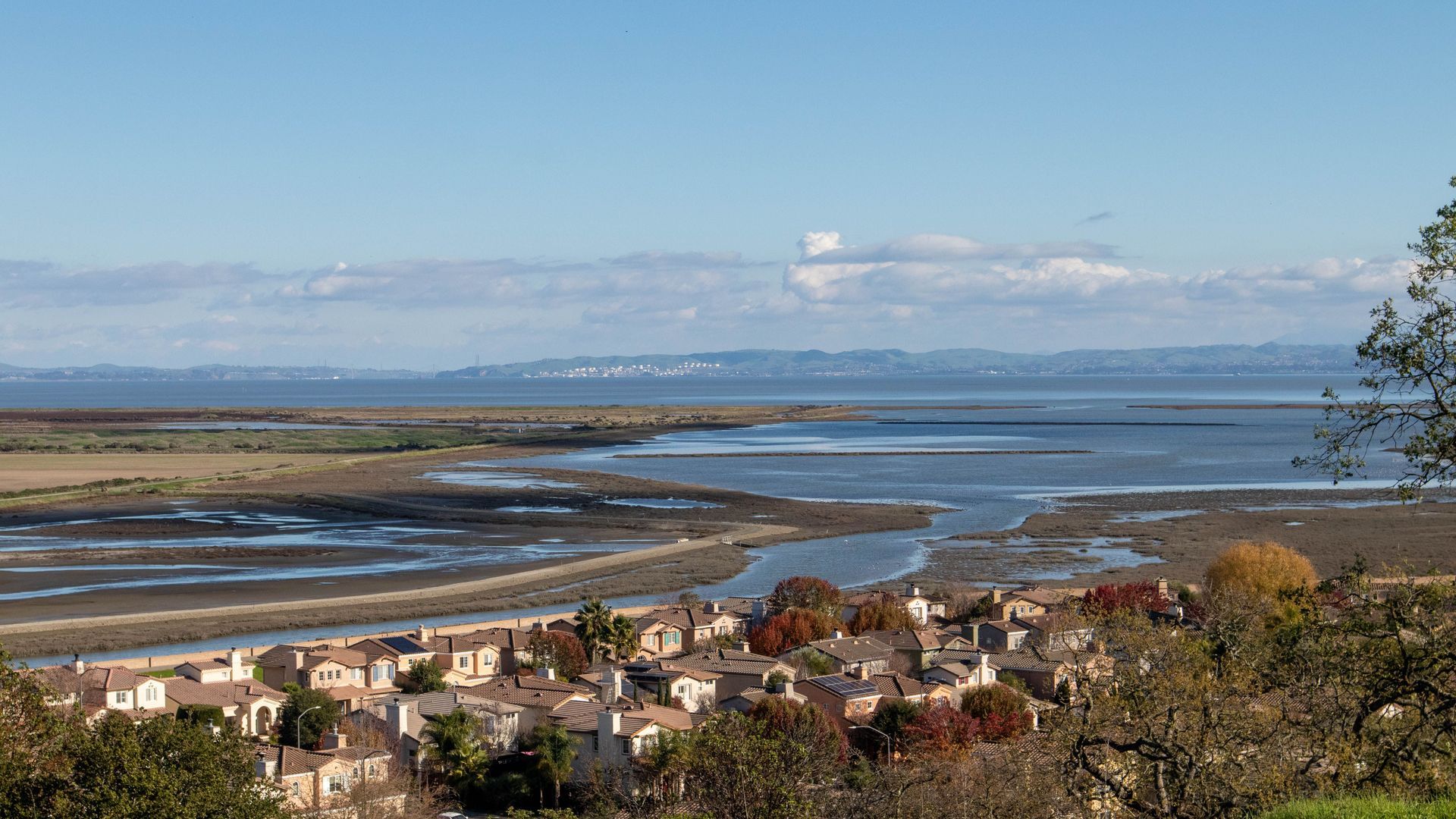 Coastal town at low tide with houses in foreground, sandy shoreline, calm water, and distant hills under a blue sky