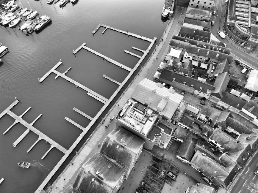 Aerial black-and-white view of a waterfront marina with empty docks beside buildings and streets.