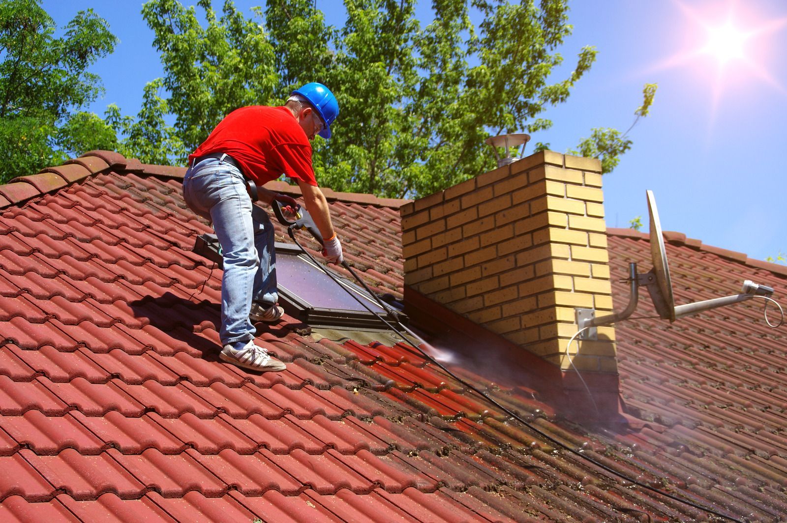 Worker pressure-washing a red-tile roof beside a brick chimney on a sunny day