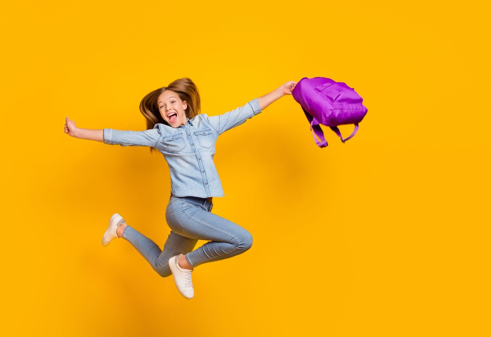 Jeune fille, son cartable violet dans les mains, sautant de joie sur fond jaune
