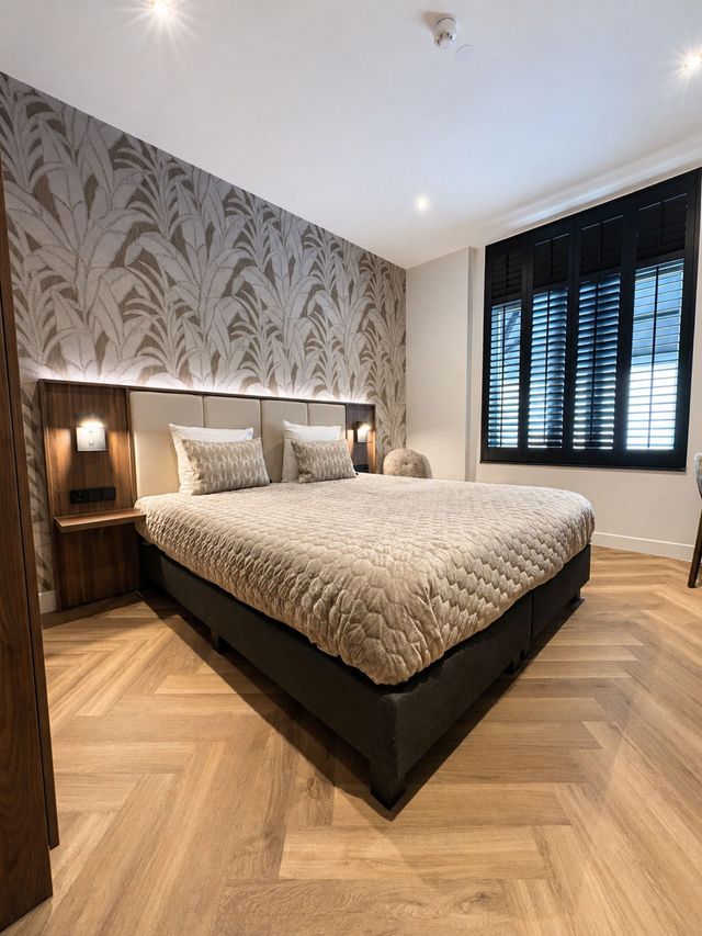 Moderne slaapkamer met behang met patroon, tweepersoonsbed, nachtlampjes en donkere raamluiken.