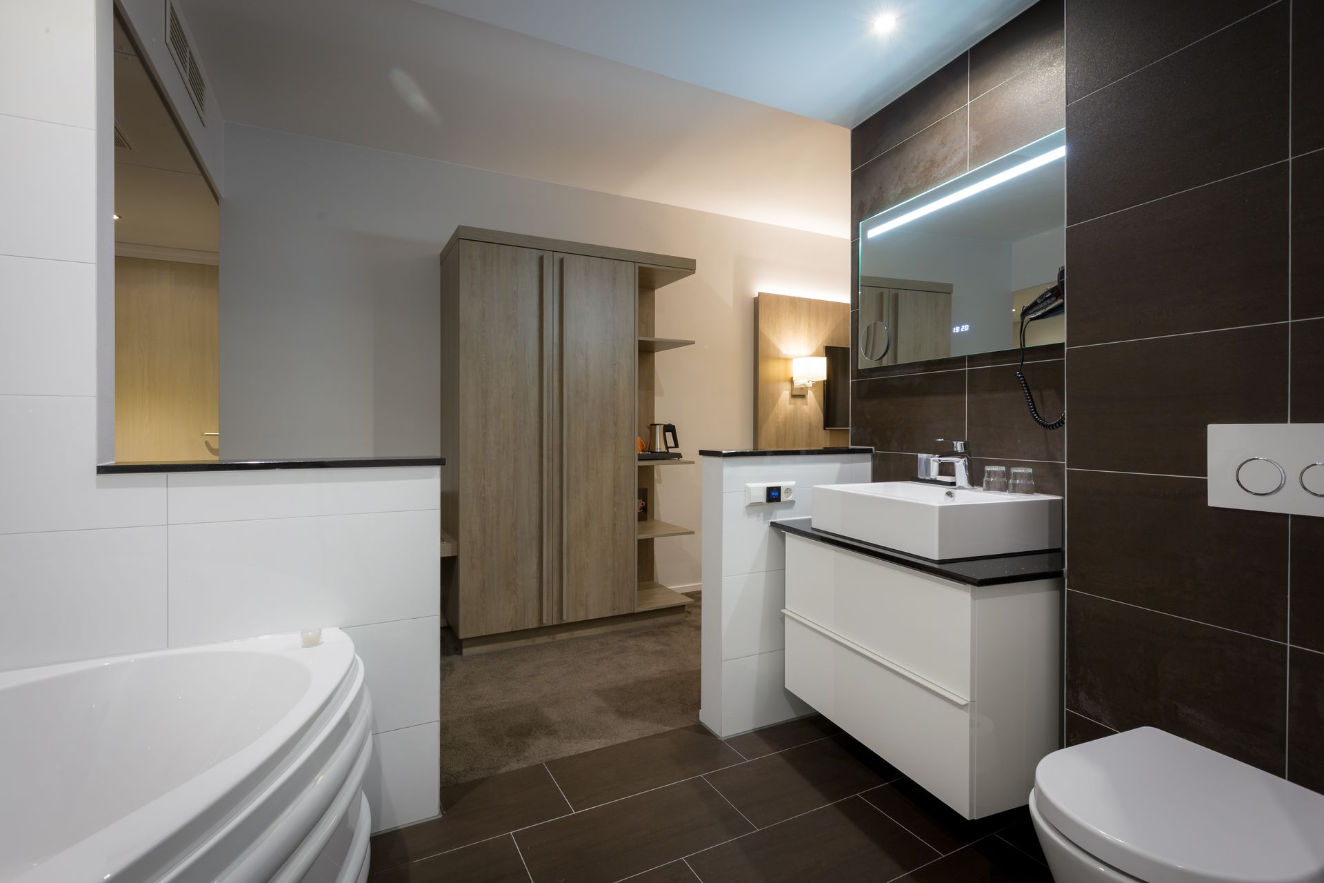Moderne badkamer met ligbad, donkere tegelvloer, witte wastafel, grote spiegel en schuifdeuren voor de douche.