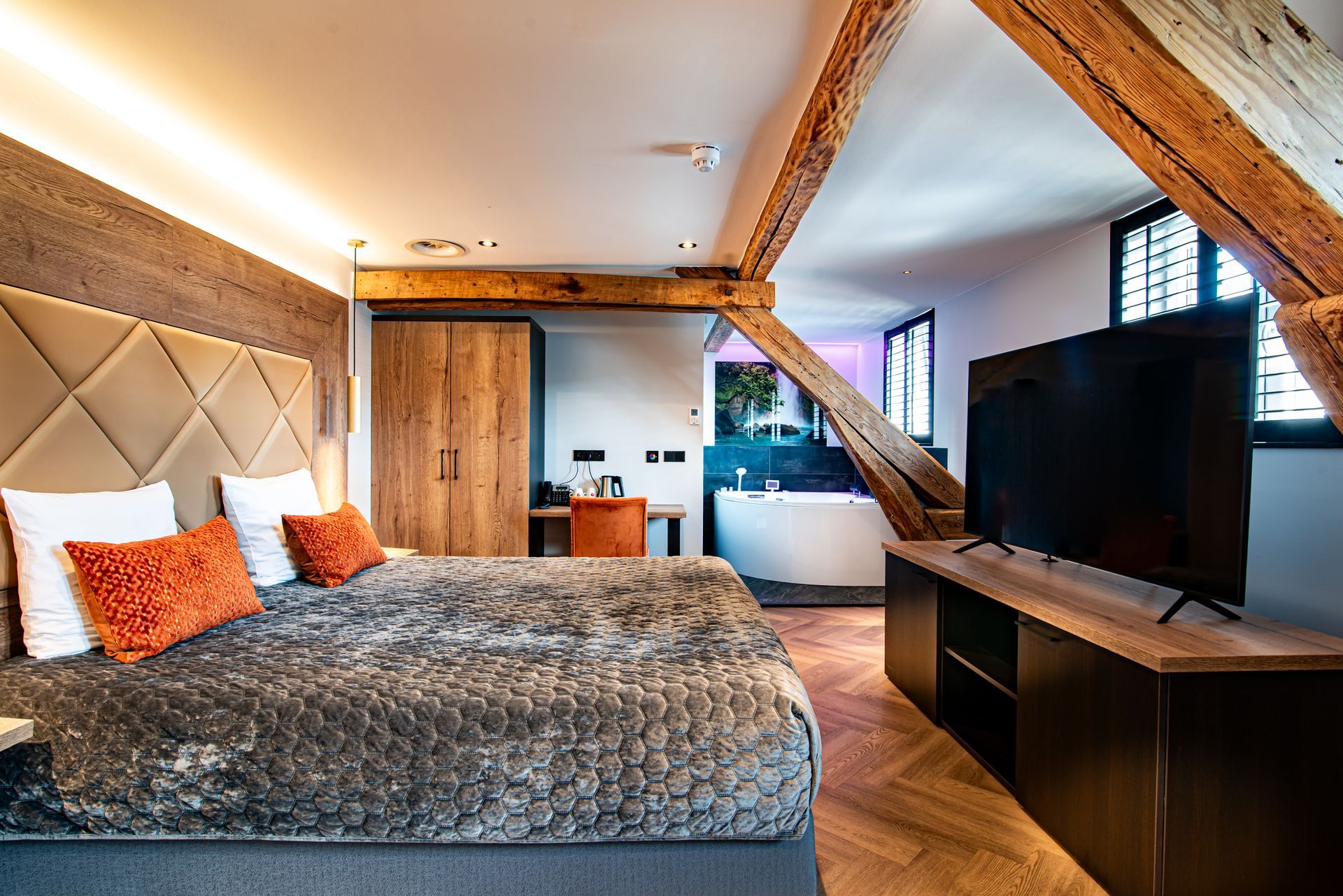 Moderne slaapkamer met een groot bed, tv, zichtbare houten balken en een open zicht op een badkamer met wastafel.
