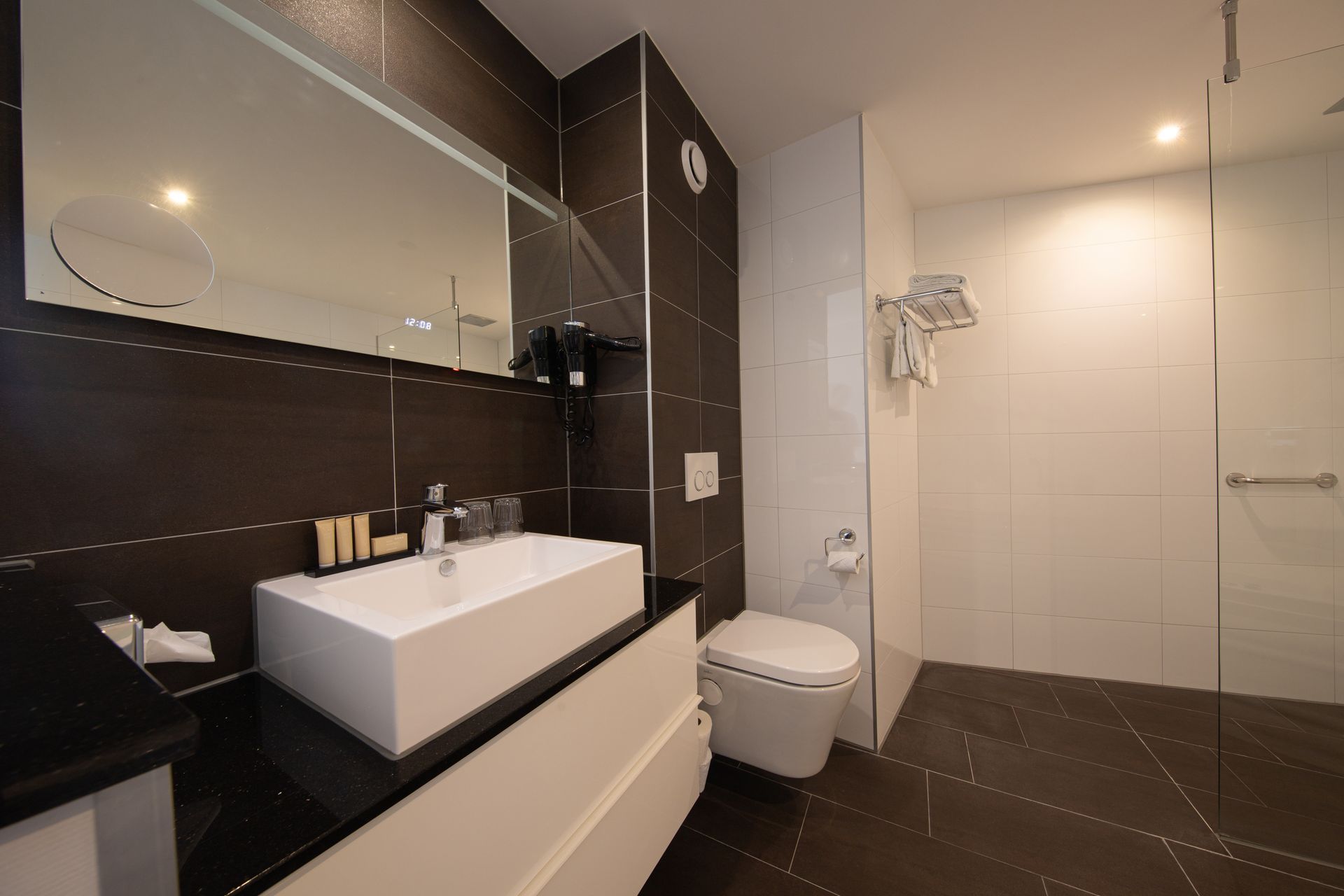 Moderne badkamer met witte wastafel, toilet, douche en donkerbruine tegelwanden en -vloer.
