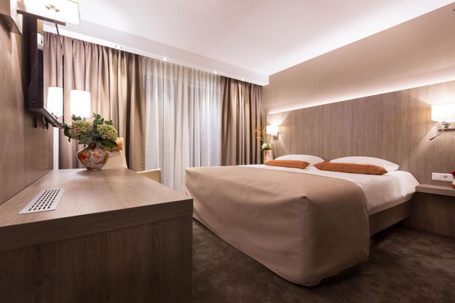 Moderne hotelkamer met een groot bed, houten lambrisering, bureau en ramen met gordijnen.