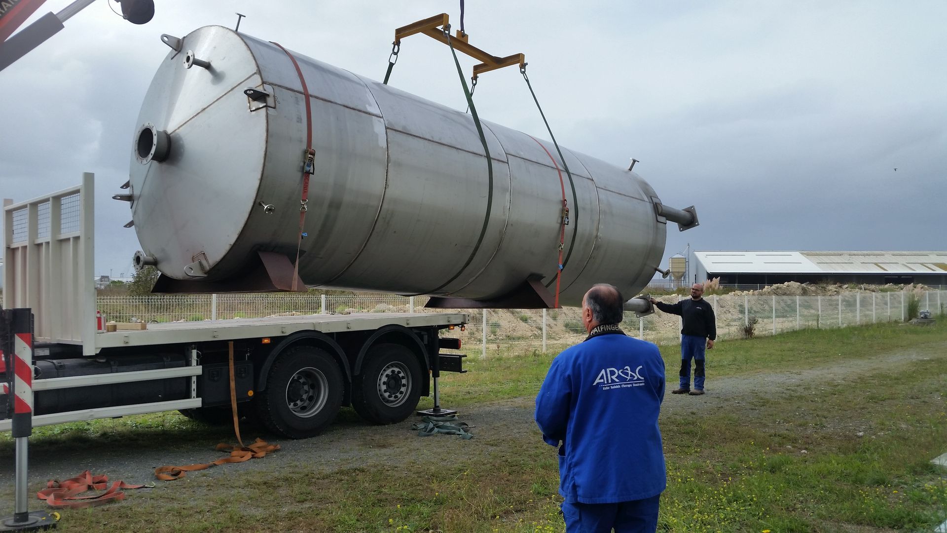 Un grand réservoir métallique est soulevé par une grue sur un camion à plateau, sous la surveillance de deux hommes.
