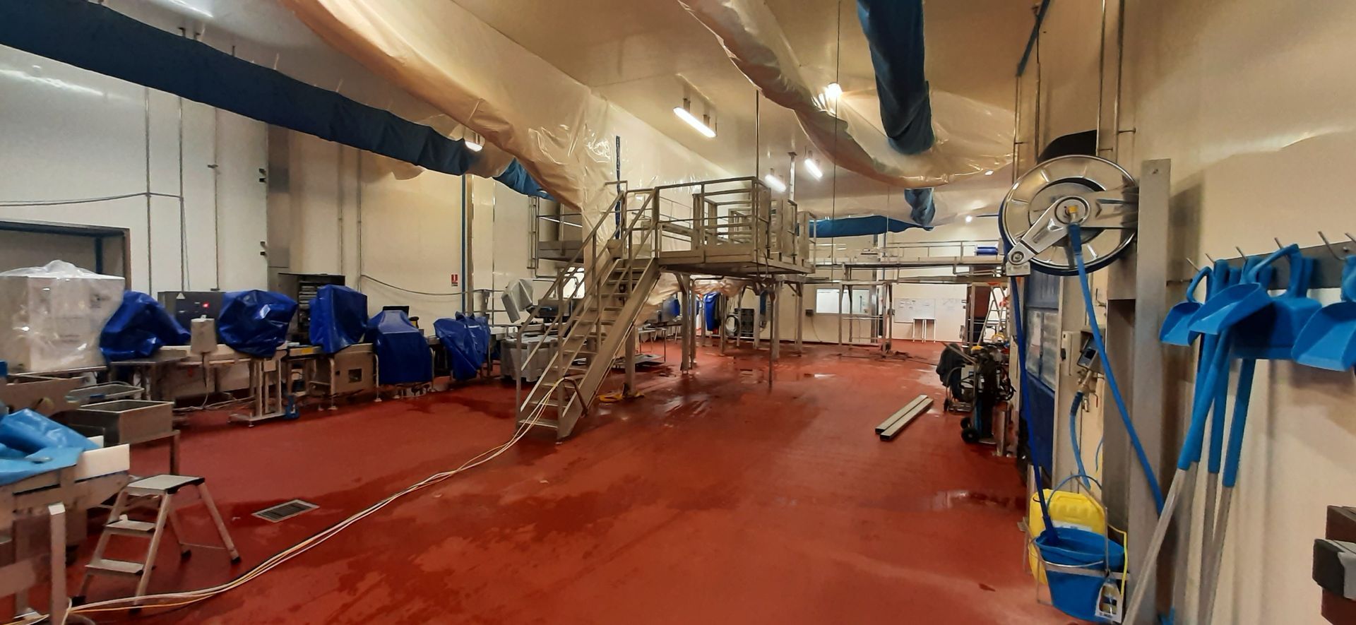 Intérieur d'une usine avec sol rouge, machines métalliques et équipements recouverts de bleu. Éclairage et tuyaux au plafond.