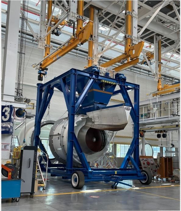 Un gros moteur à réaction soulevé par un cadre bleu et un système de grue jaune dans une usine.