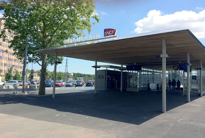 Entrée de la gare avec le logo SNCF, protégée par un auvent en bois. Voitures garées à proximité, piétons entrant.