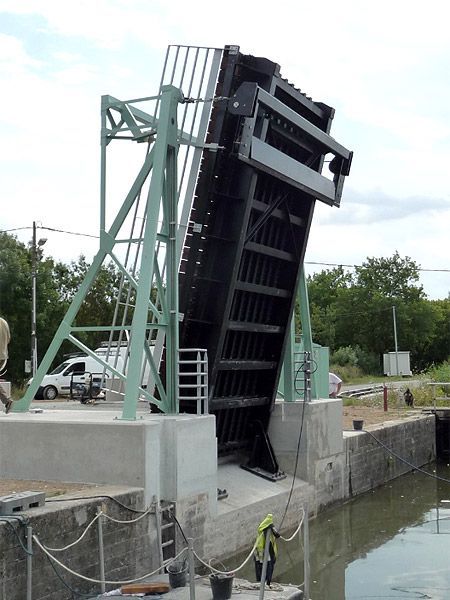 Pont-levis surélevé noir et acier au-dessus d'un canal, soutenu par une structure métallique verte ; base en béton.