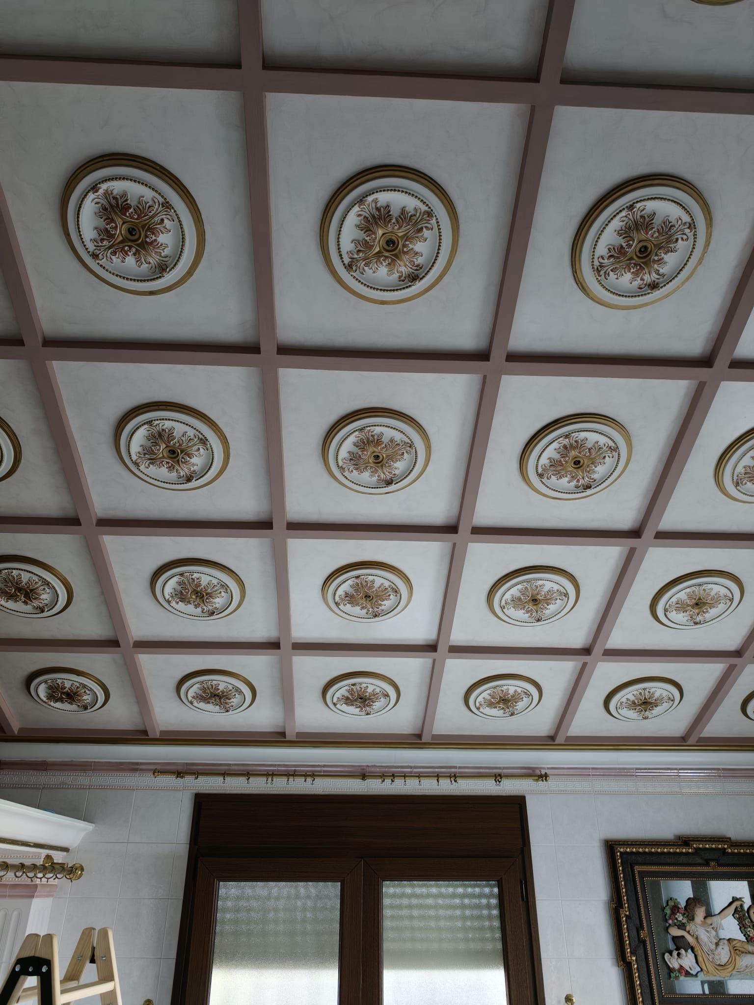 Techo blanco decorativo con medallones dorados sobre las ventanas de una habitación y arte en la pared.