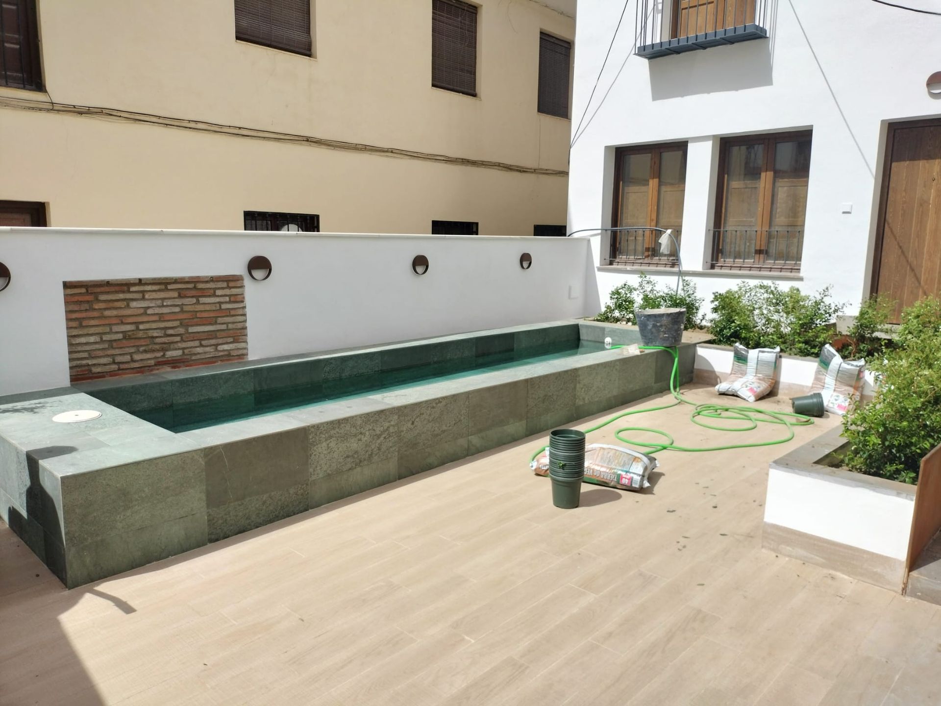 Pequeño patio con una piscina rectangular alargada, azulejos beige y paredes blancas del apartamento con plantas.