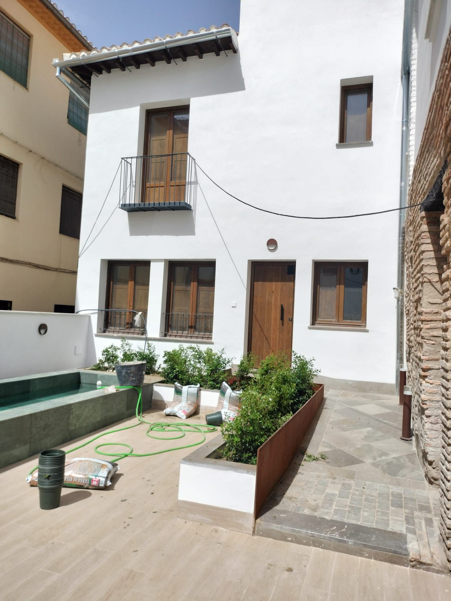 Patio blanco con una casa de dos plantas, puertas y ventanas de madera, plantas en macetas y una pequeña piscina.