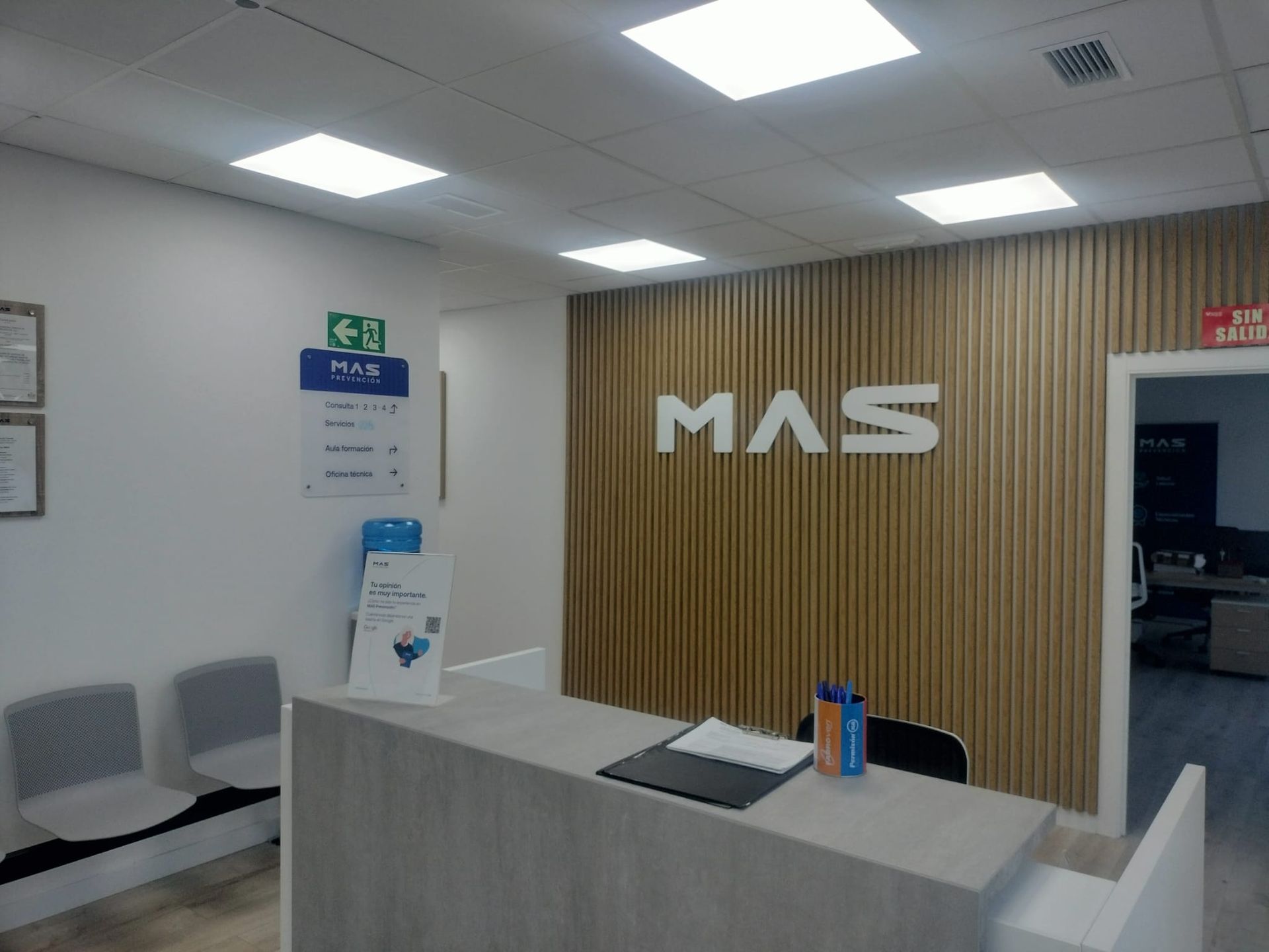 Mostrador de recepción de oficina moderno con letrero “MAS”, sillas grises, pared de madera y luces de techo brillantes.