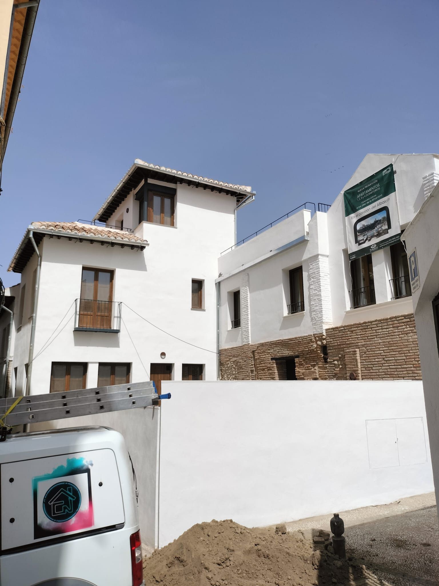 Casas blancas de estuco con tejados de tejas detrás de un alto muro bajo un cielo azul despejado.