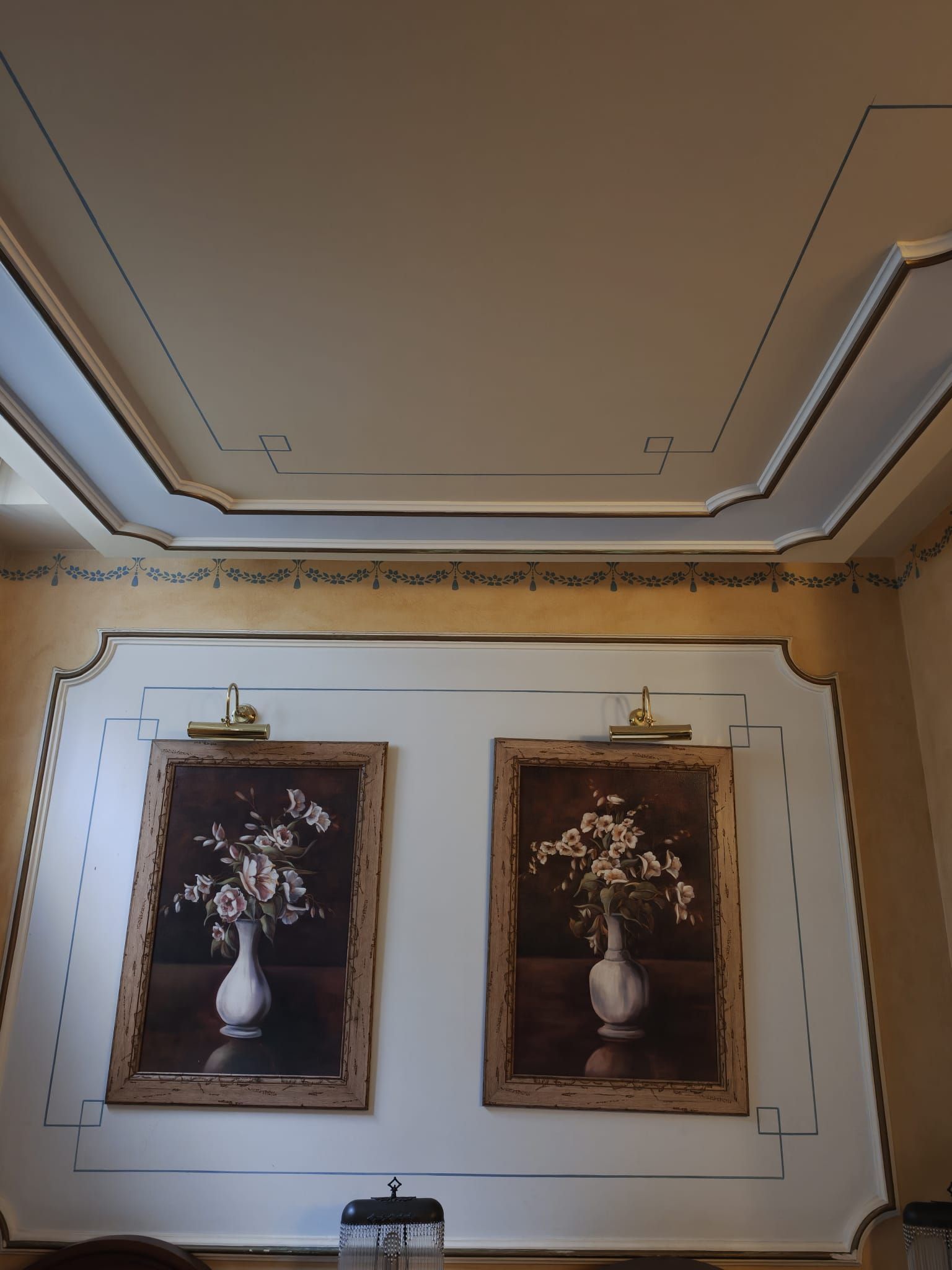 Techo y pared con molduras ornamentadas, con dos cuadros enmarcados de bodegones florales sobre una pequeña mesa.