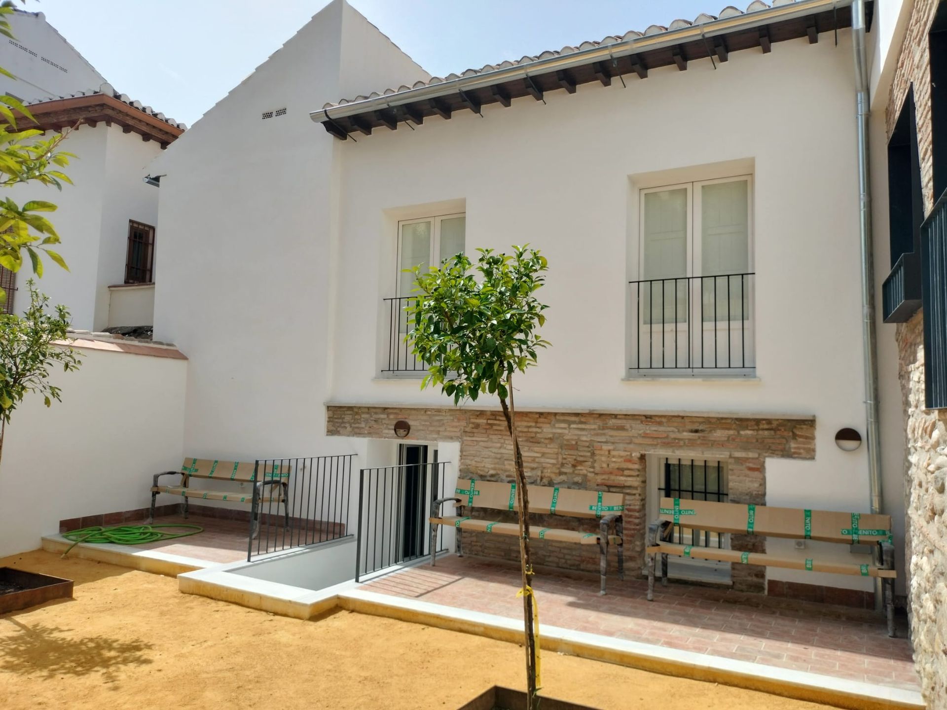 Patio blanco con un pequeño árbol, bancos y ventanas en la fachada de estuco de una casa.