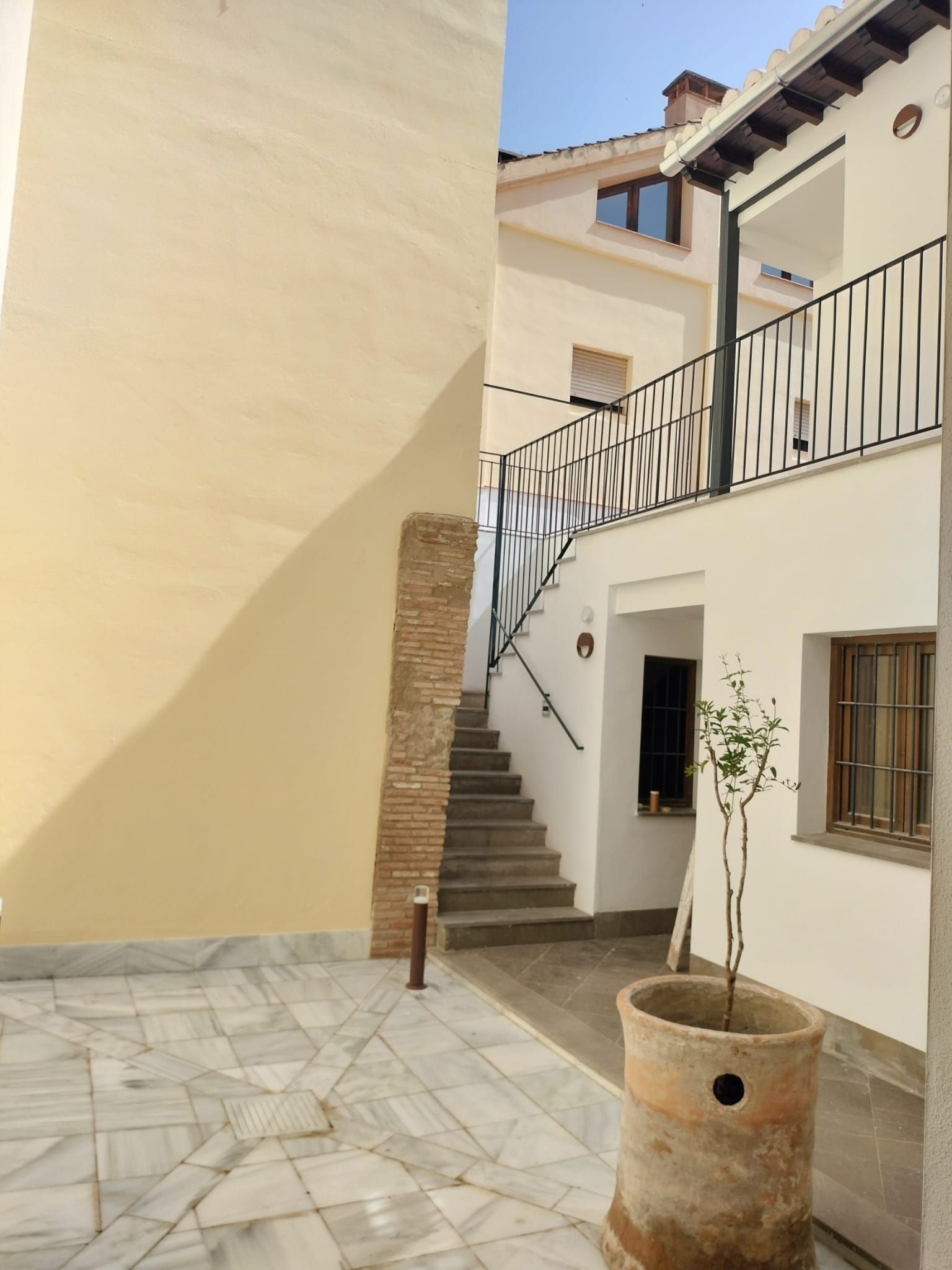 Patio soleado con escaleras de piedra, edificios blancos, balcón de hierro forjado y un árbol en maceta en primer plano.
