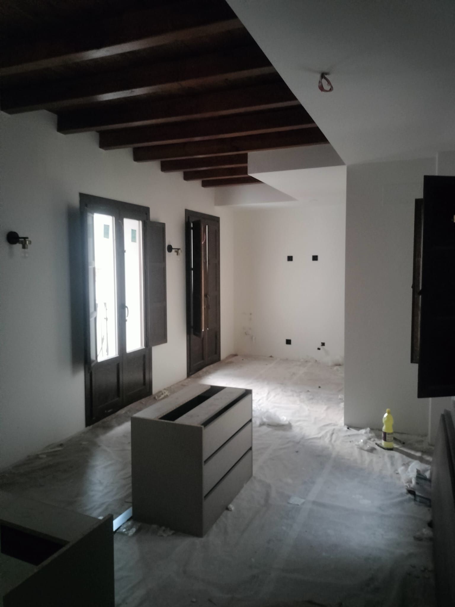 Habitación sin terminar con paredes blancas, vigas a la vista y materiales de construcción en el suelo.