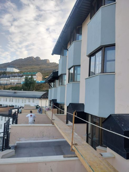 Pasarela junto a un edificio azul con montañas al fondo y una persona vestida de blanco abajo.
