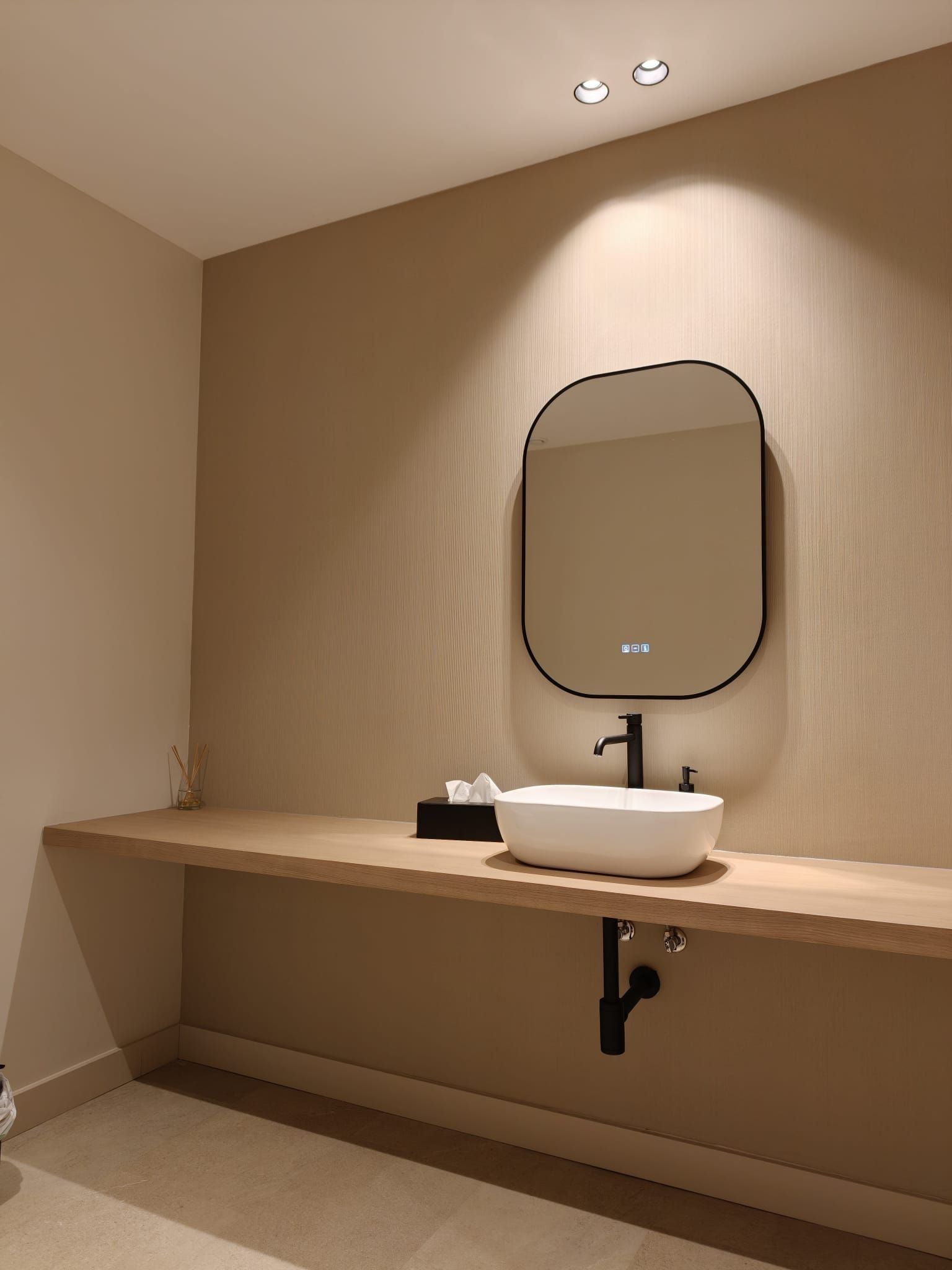 Baño moderno de color beige con tocador de madera, espejo ovalado y lavabo blanco sobre encimera bajo iluminación cálida.