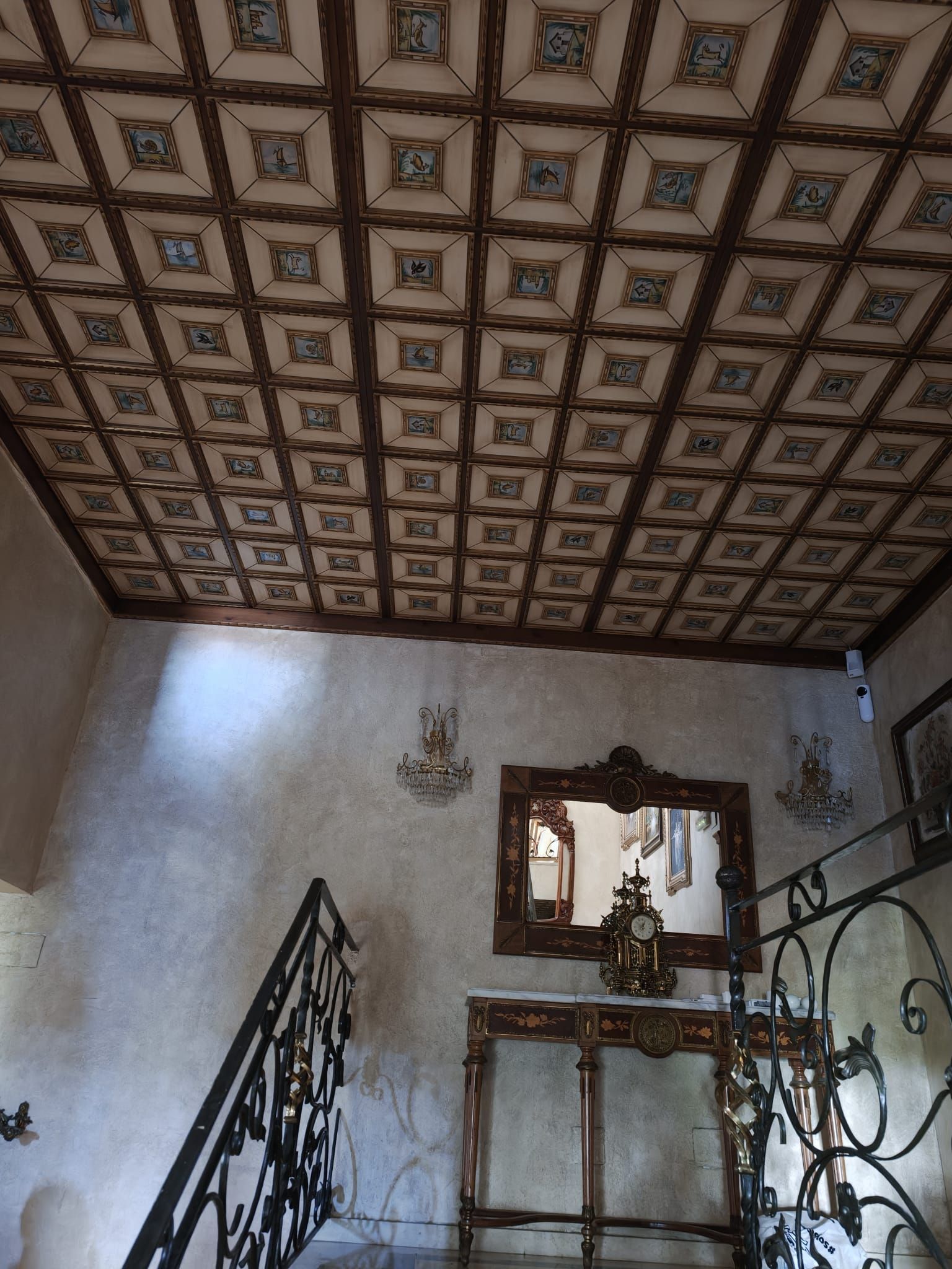 Rellano de escalera con techo decorado con elaborados motivos, espejo de pared y barandillas de hierro forjado decorativas.