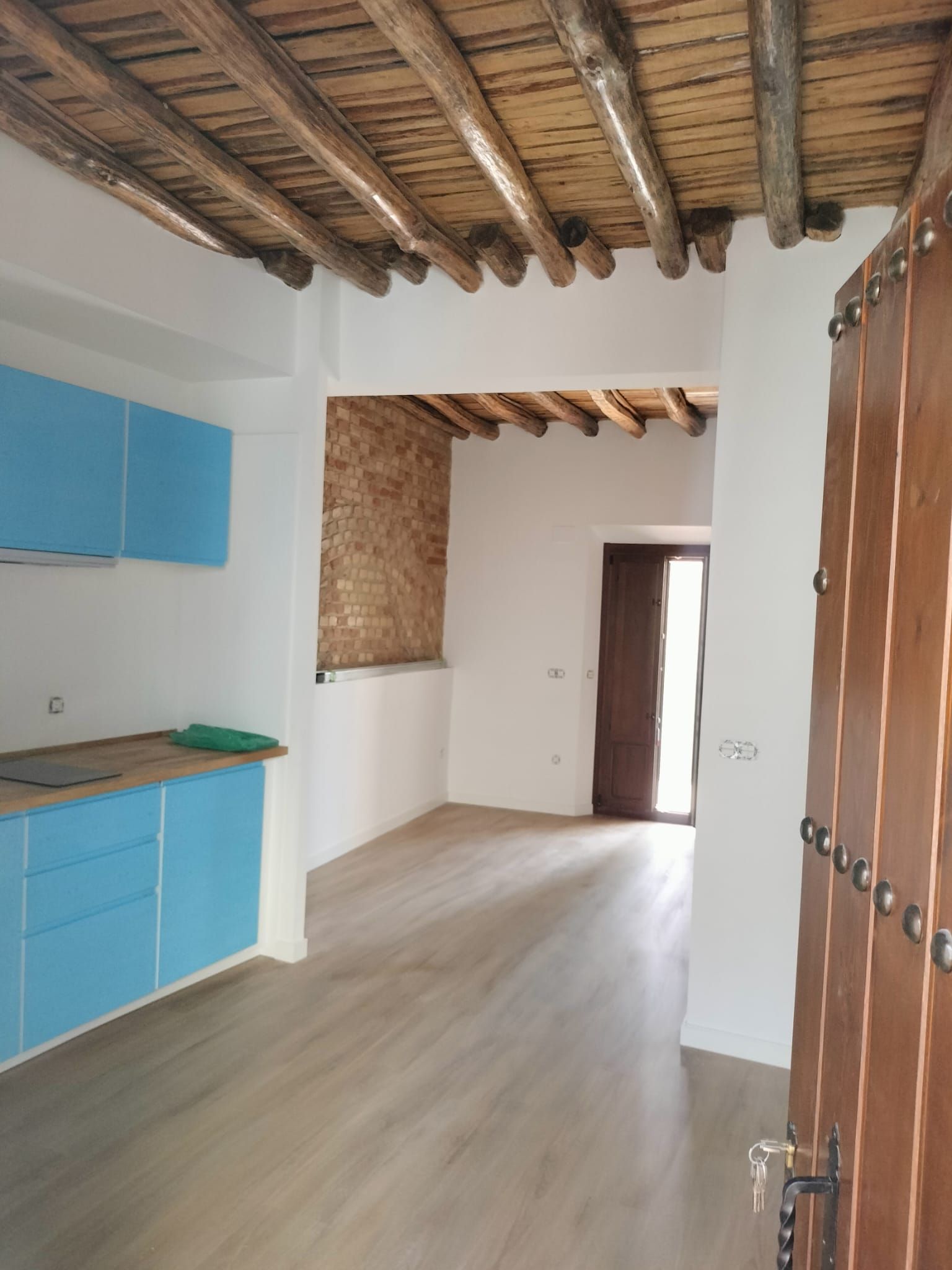 Cocina rústica estrecha con armarios azules, vigas de madera a la vista y una puerta que da a una habitación luminosa.