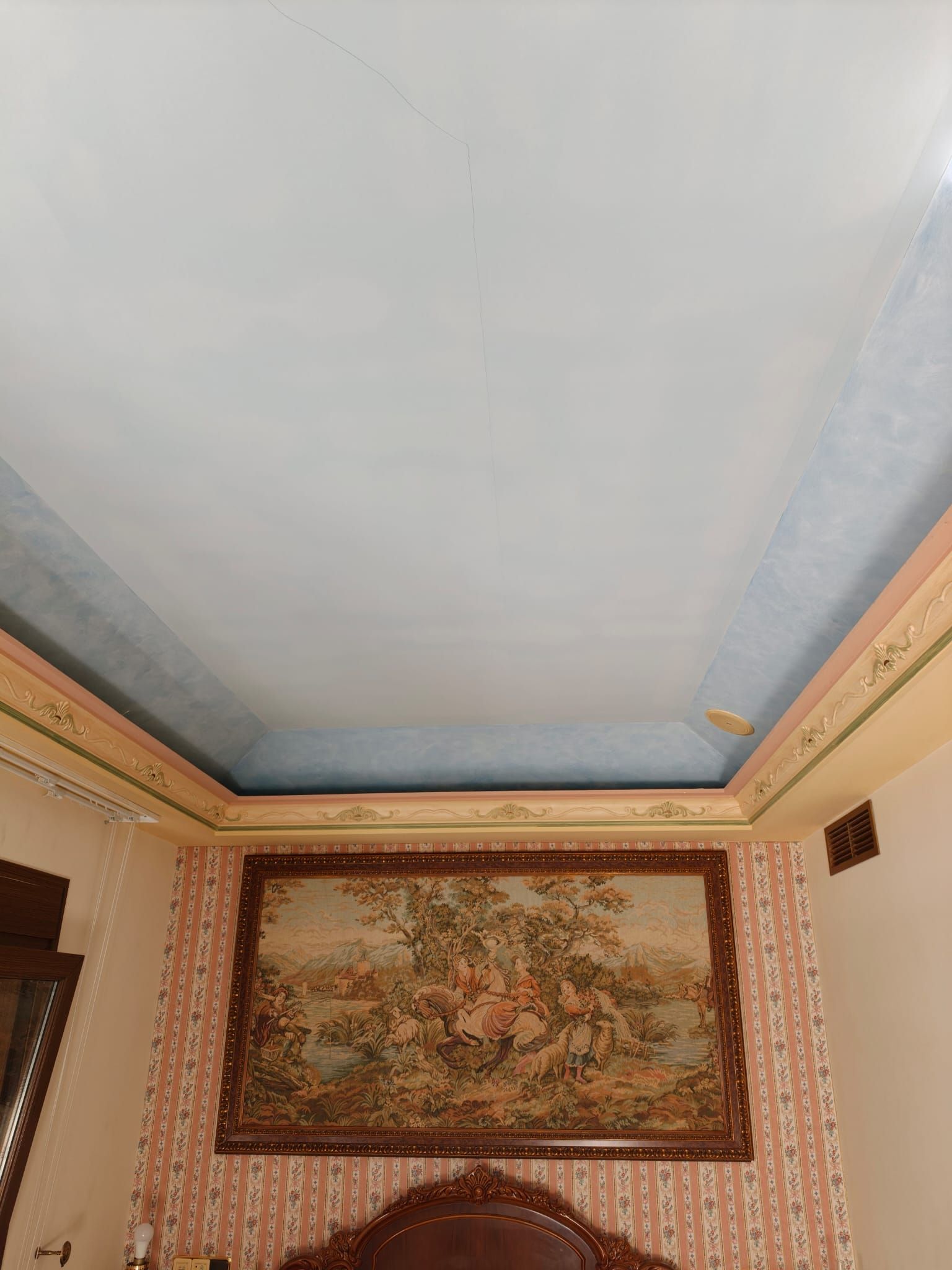 Rincón de una habitación con techo azul, papel tapiz estampado y un tapiz enmarcado en la pared.