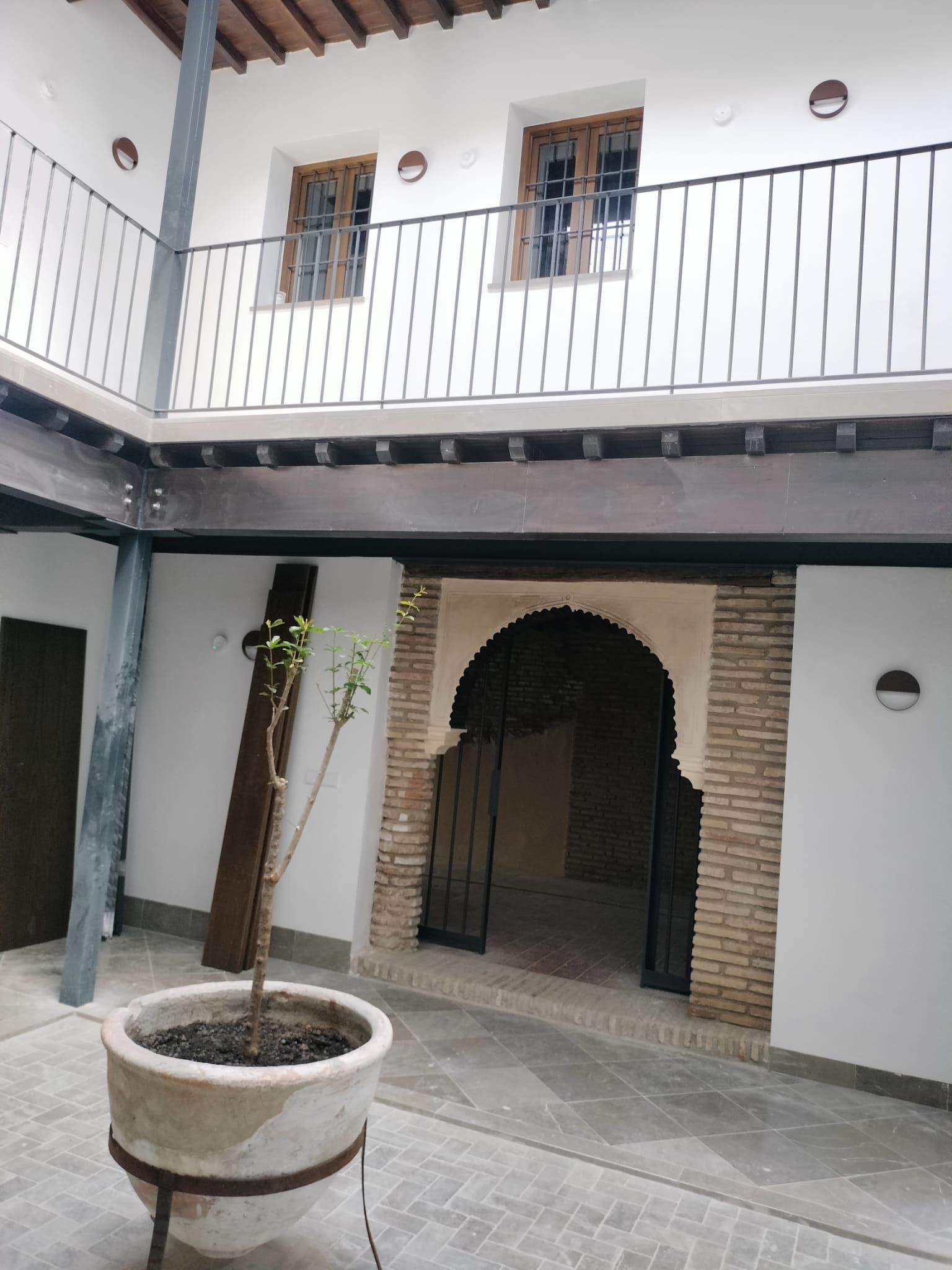 Patio interior con paredes blancas, puerta arqueada, balcón superior y un gran árbol en maceta.