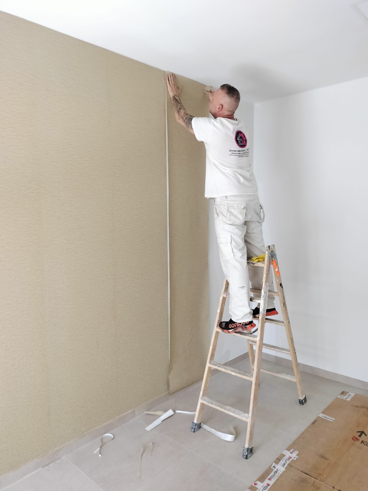 Un trabajador subido a una escalera pinta una pared beige en una habitación con protección de plástico en el suelo.