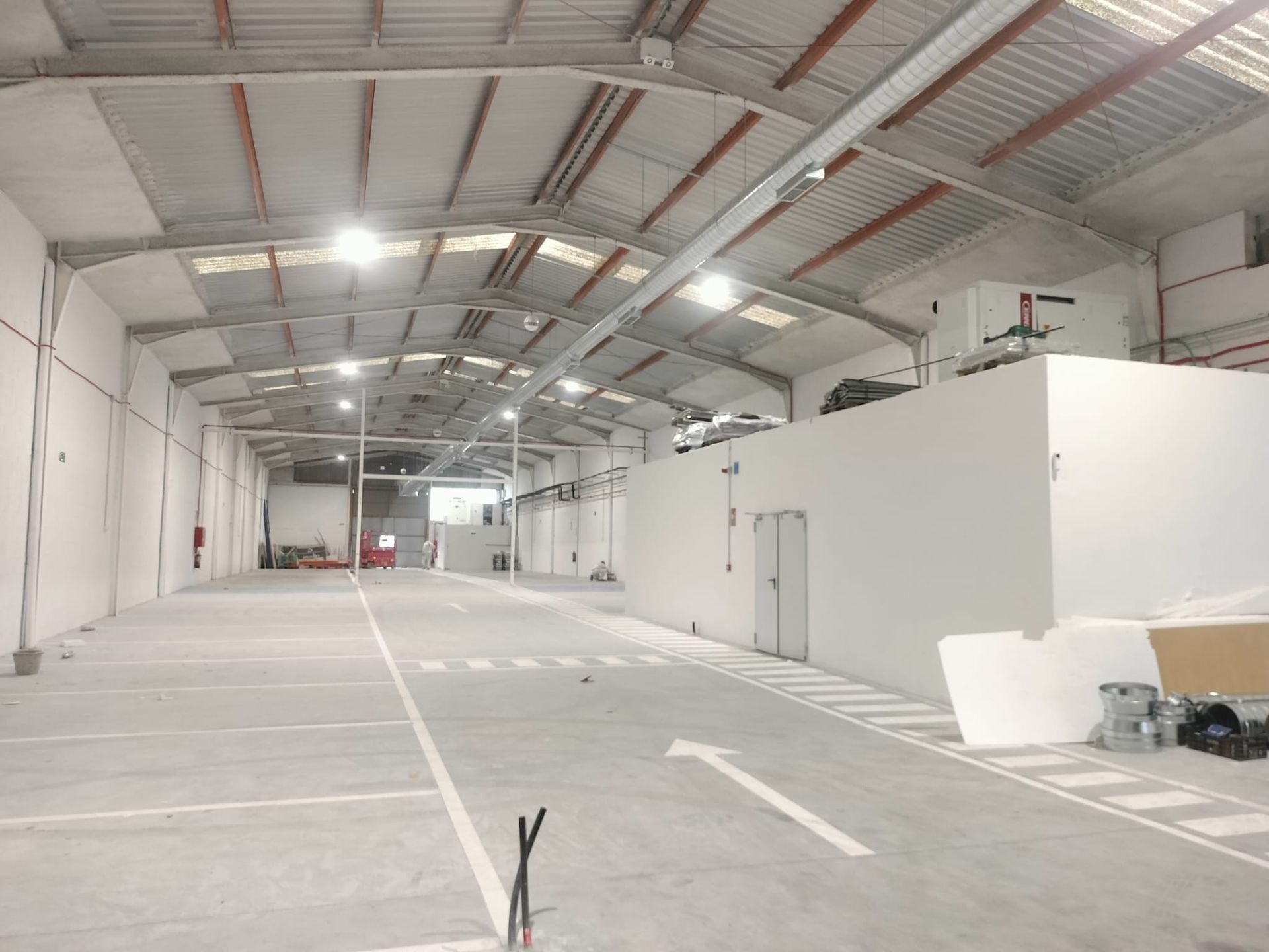 Nave industrial vacía con paredes blancas, techo alto de metal y carriles de suelo de hormigón señalizados.