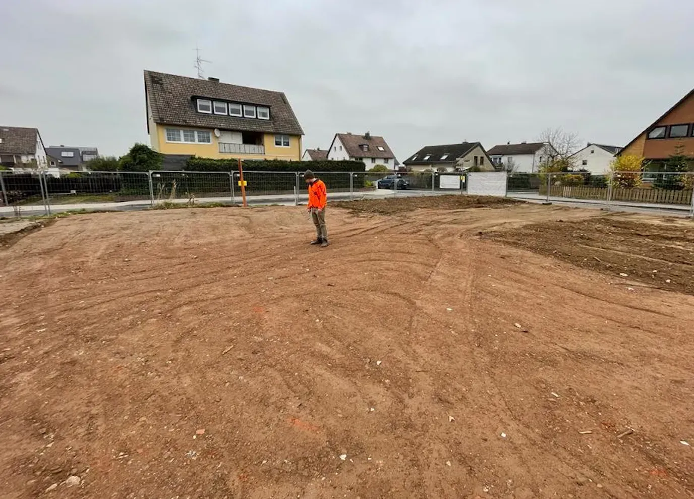 Eine Person in orangefarbener Weste steht auf einem großen unbebauten Grundstück; im Hintergrund sind Häuser und ein bewölkter Himmel zu sehen.