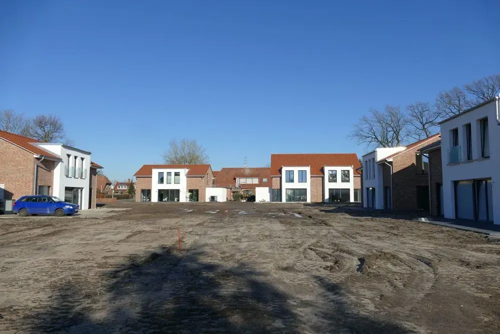 Eine Reihe moderner Stadthäuser umgibt einen kahlen, unbefestigten Innenhof unter klarem, blauem Himmel