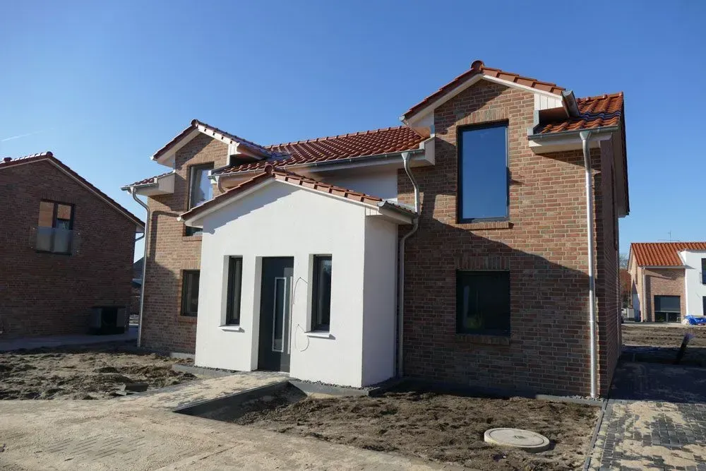 Neues Backsteinhaus mit weißem Vorderanbau und rotem Ziegeldach auf einem unbefestigten Grundstück