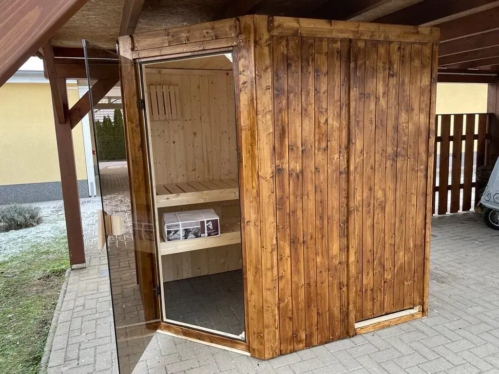 Referenzbild: Kleine Sauna aus dunklem Holz unter einem erweiterten Carport