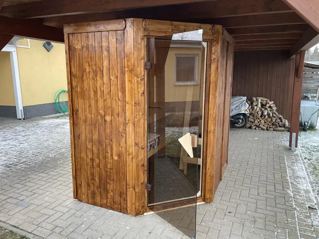 Referenzbild: Kleine Sauna aus dunklem Holz unter einem erweiterten Carport
