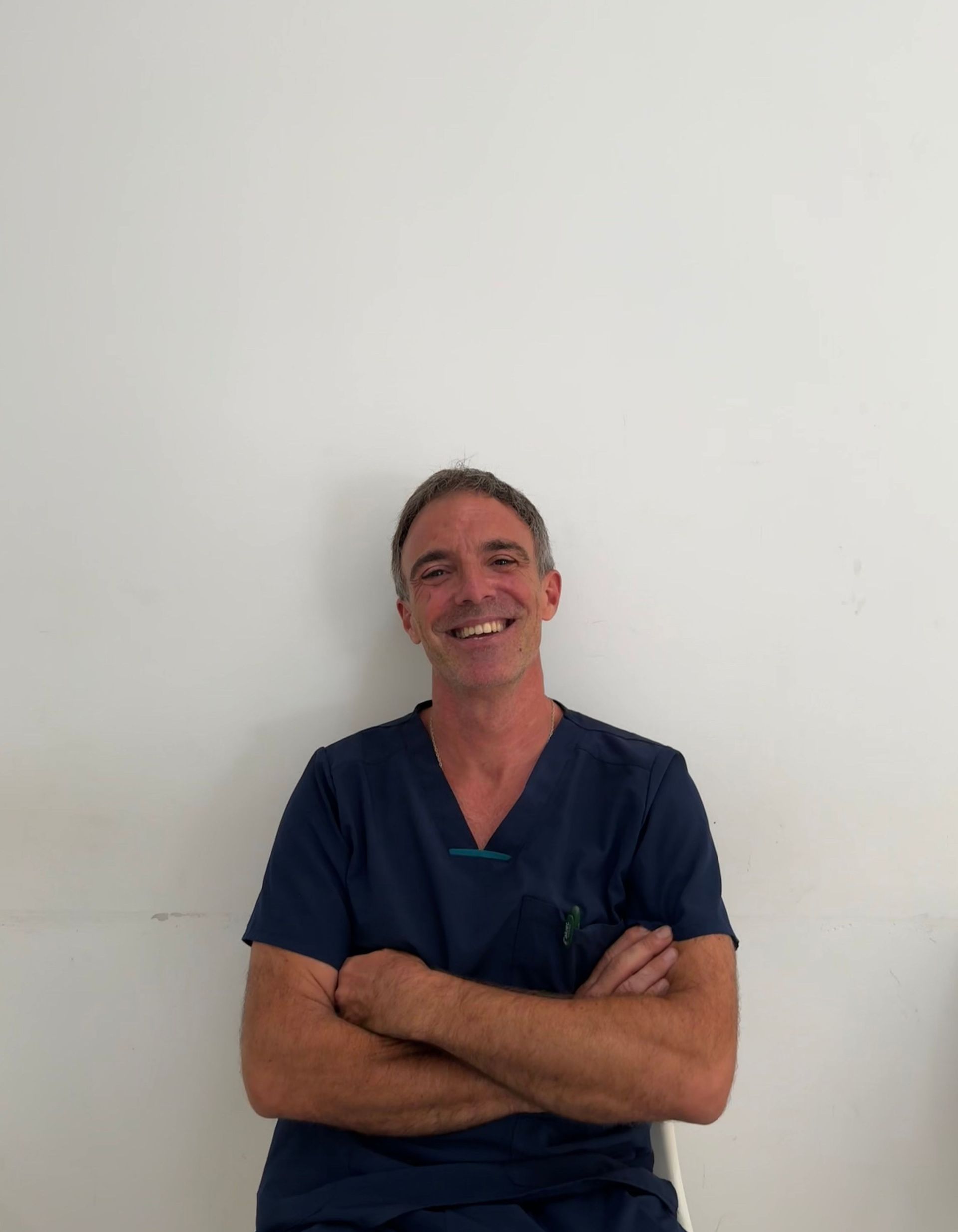 Un hombre con uniforme médico está sentado en una silla con los brazos cruzados y sonriendo.
