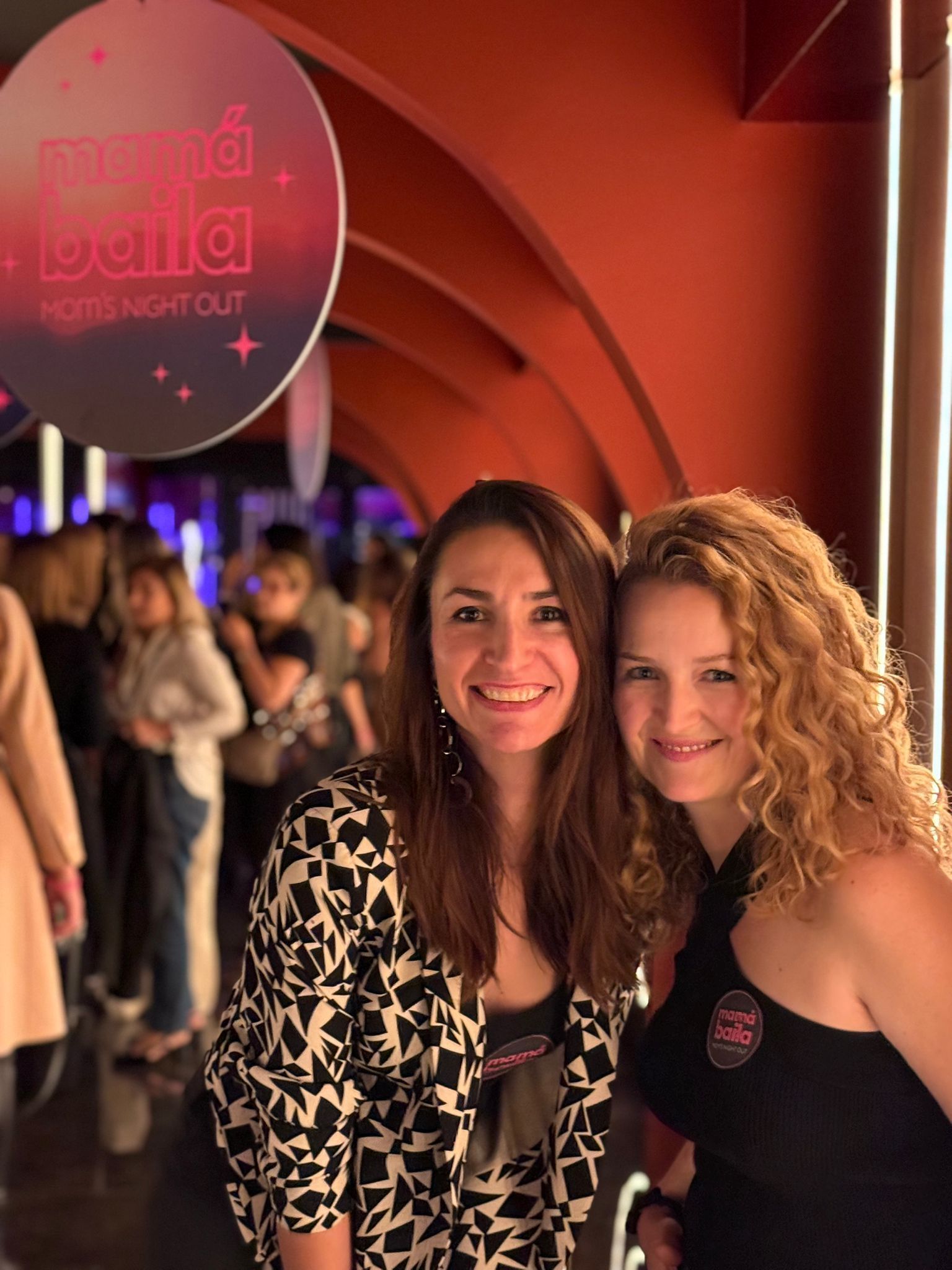 Dos mujeres sonrientes posan dentro de un local; el cartel de arriba dice 