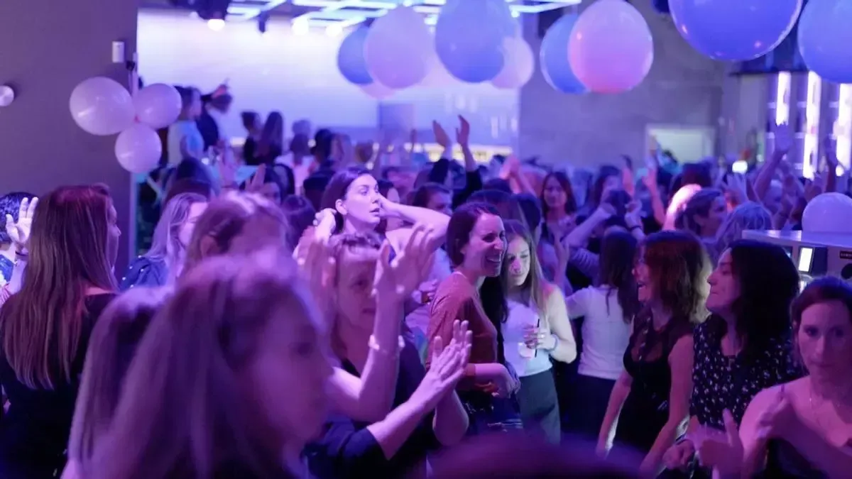 Gente bailando en un club bajo globos azules y blancos, iluminación violeta.