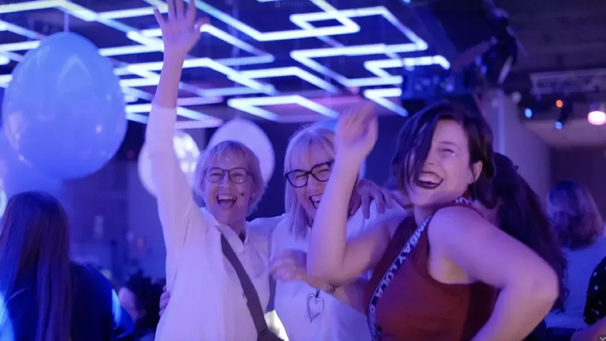 Tres personas bailando en una fiesta bajo luces azules; uno con los brazos en alto, el otro sonriendo.