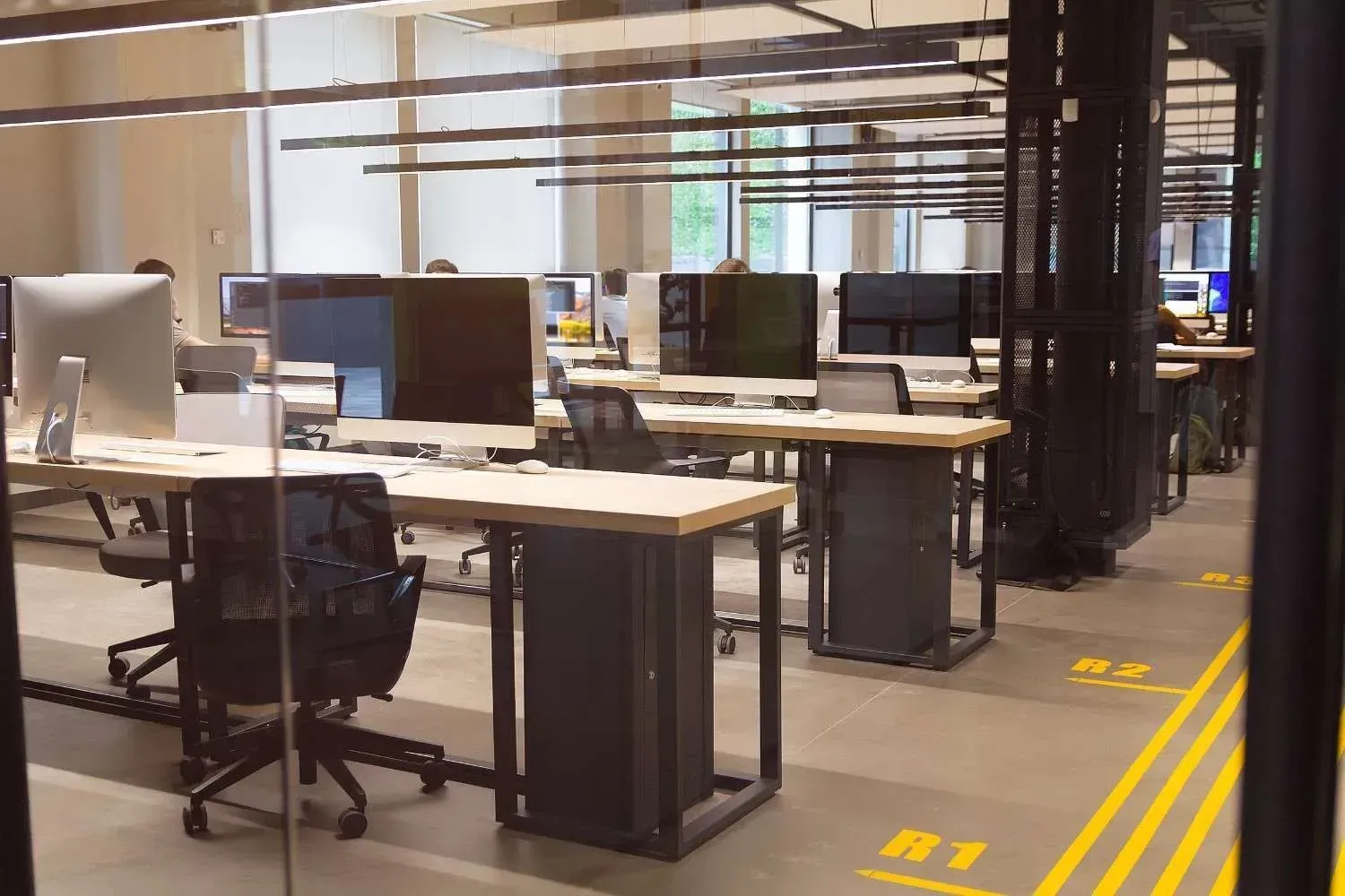 Oficina moderna y vacía con escritorios, monitores y sillas de oficina bajo luces cenitales brillantes.