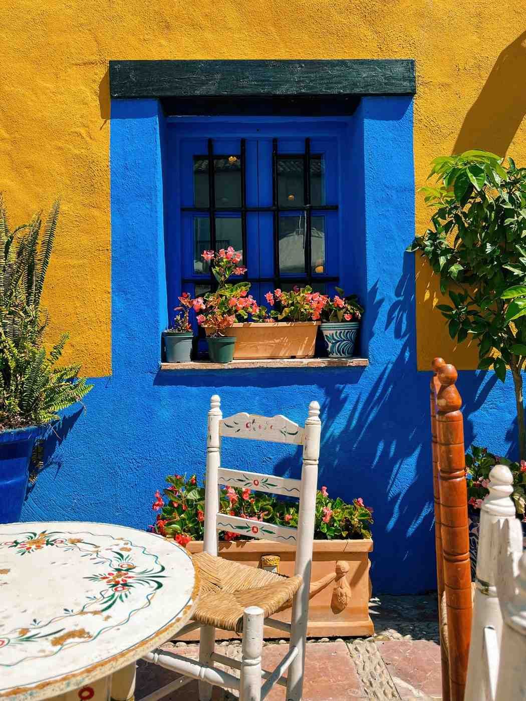 Patio colorido con ventana azul, pared amarilla, jardineras y sillas blancas junto a una mesa.