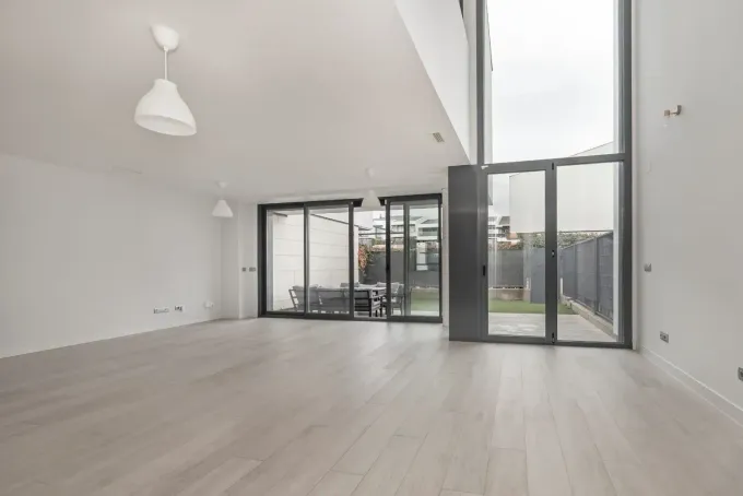 Luminoso salón moderno y vacío con suelos de madera clara, paredes blancas y grandes puertas de cristal que dan a un patio.