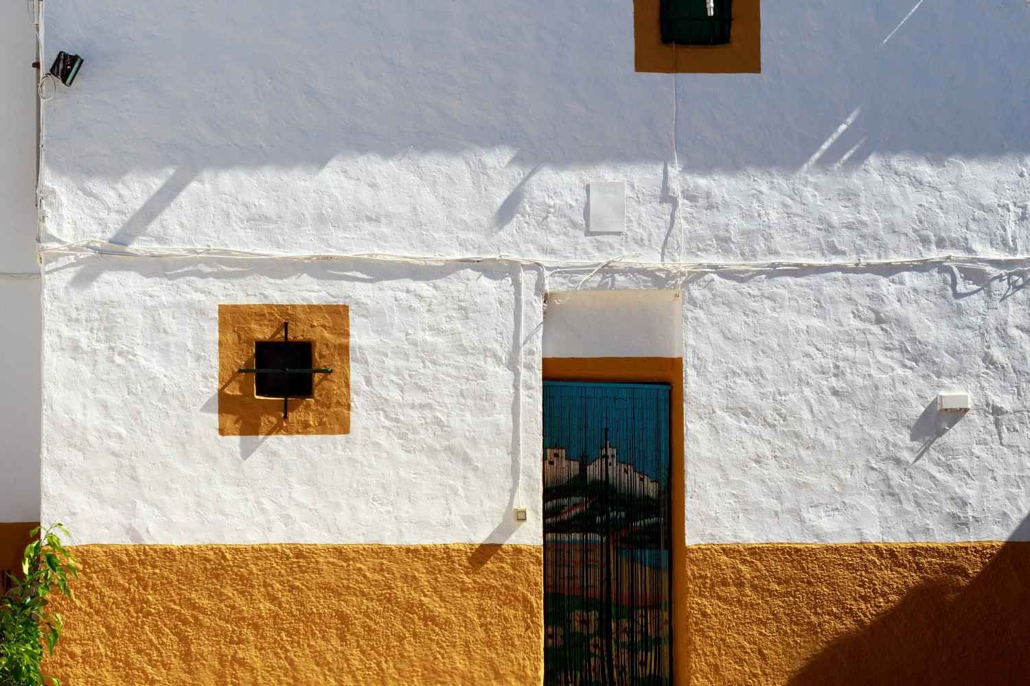 Pared de estuco blanco con base naranja, ventana pequeña y puerta central con cortina azul.