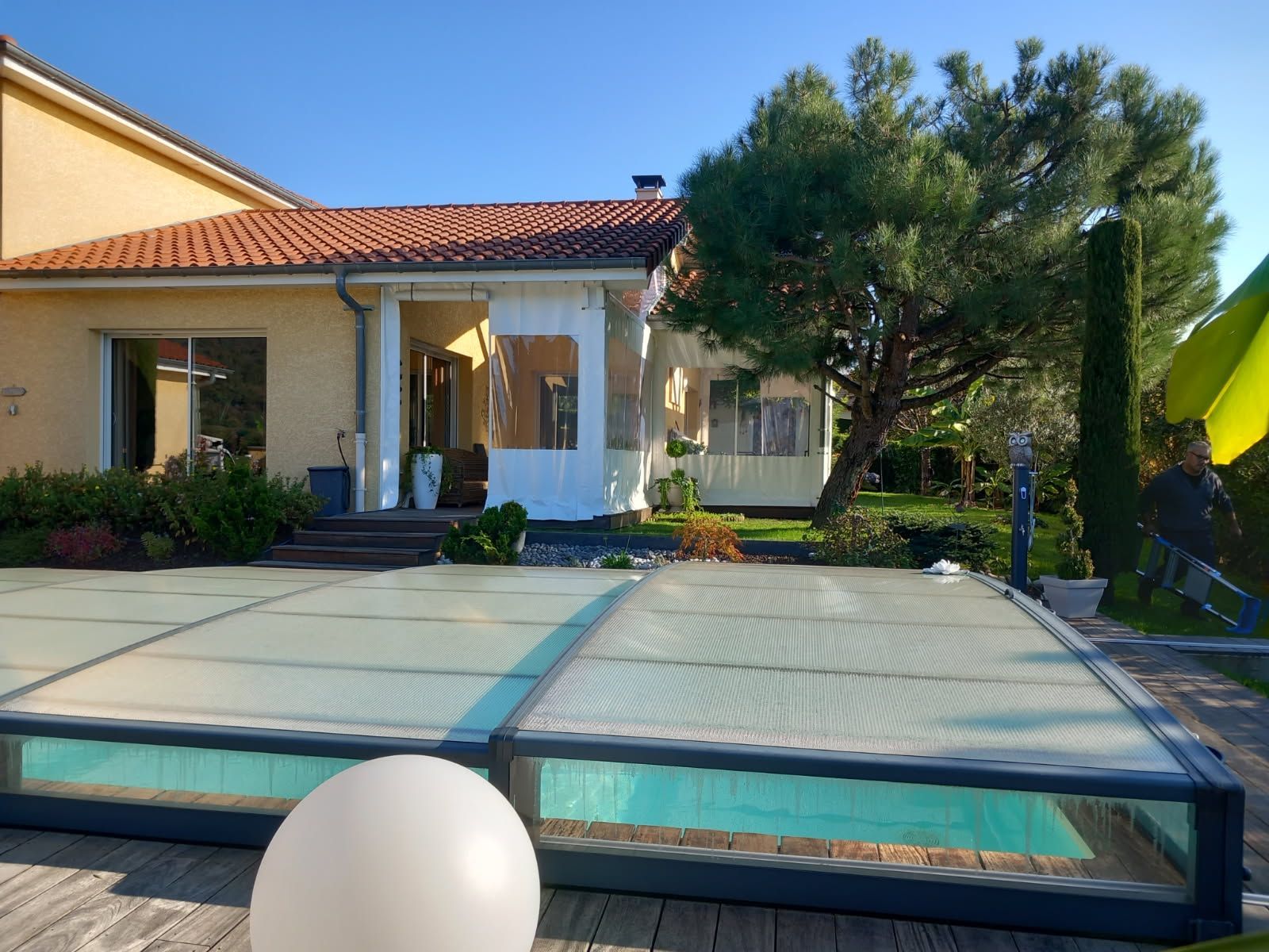 Une piscine couverte dans un jardin avec une maison jaune et des arbres.