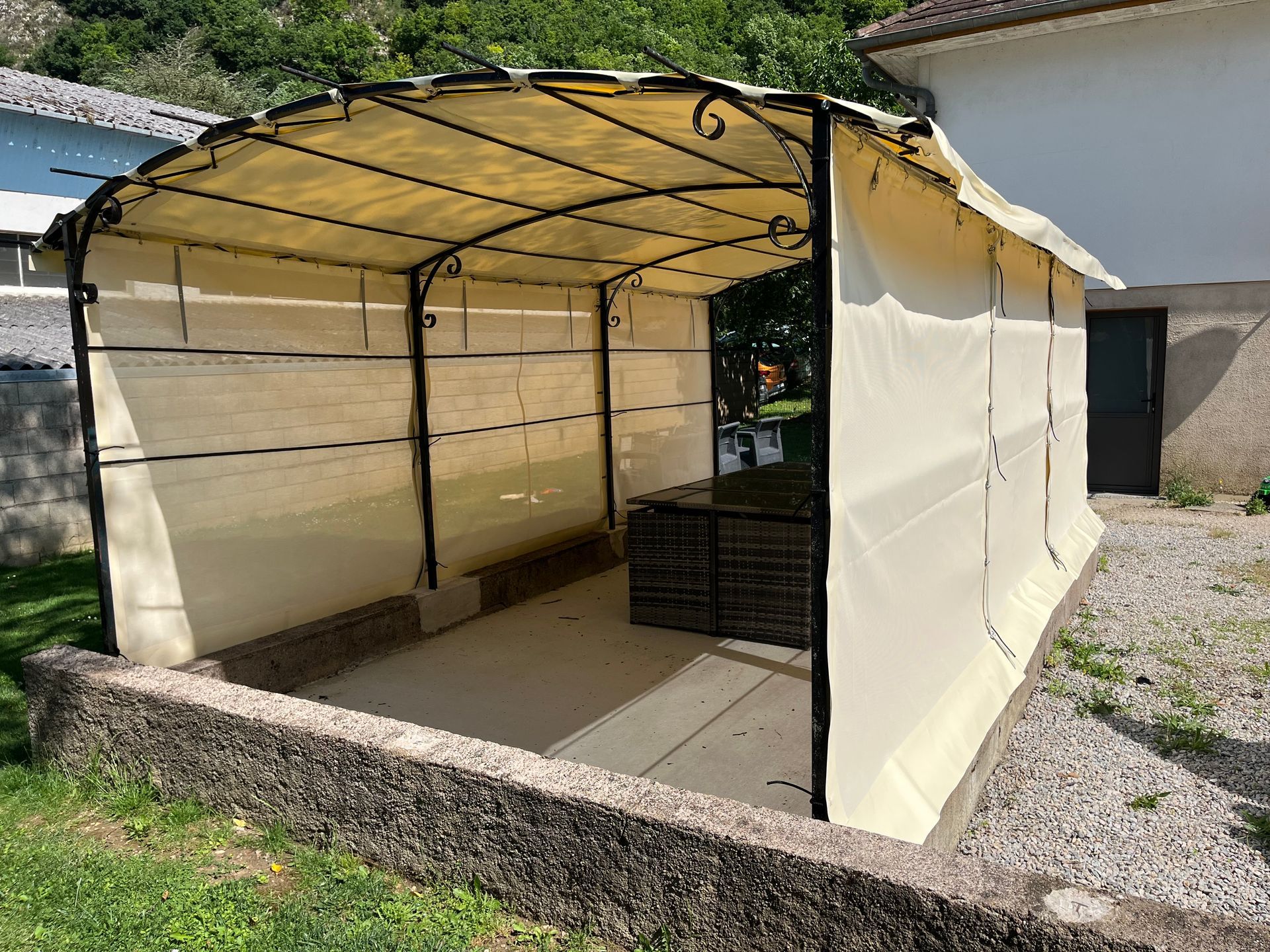 Abri de jardin beige à structure métallique noire, abritant du mobilier de terrasse, installé sur une terrasse en béton.